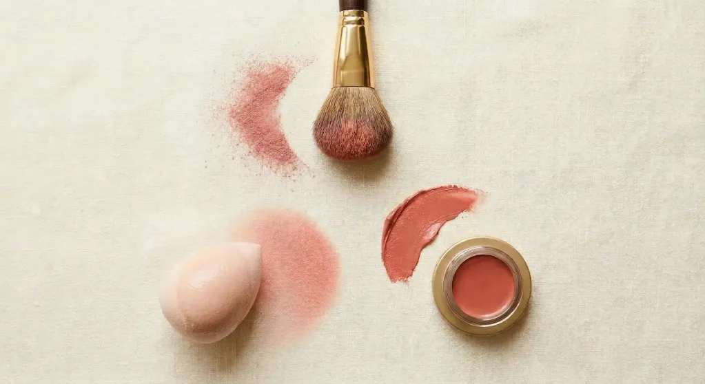 Disposition triangulaire d'un pinceau à blush duveteux, d'une éponge à maquillage humide et d'une main avec un échantillon de blush crème sur le bout du doigt, sur une surface crème chaude